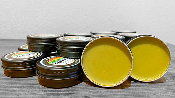 View of black Ph other on a wooden table with twoappy's tins stacked on top of each tins resting on their sides with the lid off exposing a view of the product which is a yellow-green color